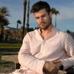 young hispanic male wearing a pink shirt and posing on the beach near palm trees